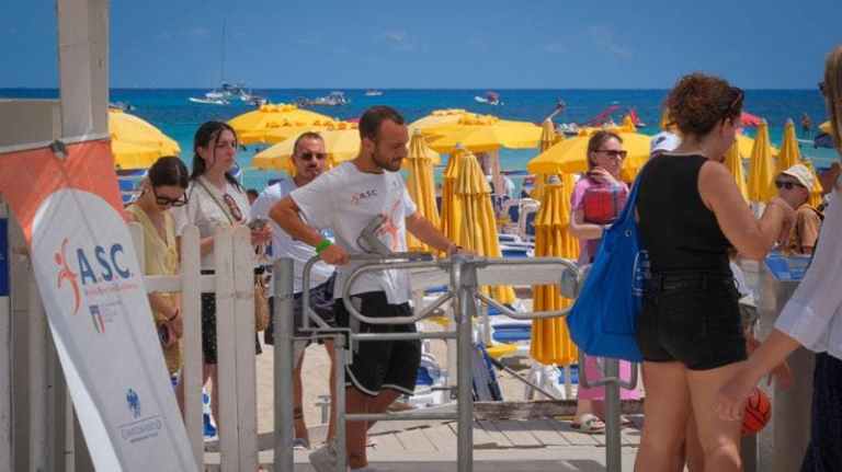 Persone passano attraverso i tornelli della spiaggia di Mondello con ombrelloni gialli sullo sfondo in una giornata estiva.