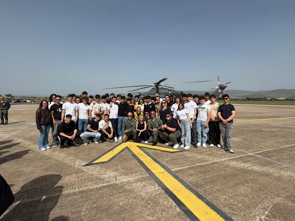 Un gruppo di studenti in posa davanti a un elicottero su un campo di aviazione, con un ufficiale militare in uniforme.